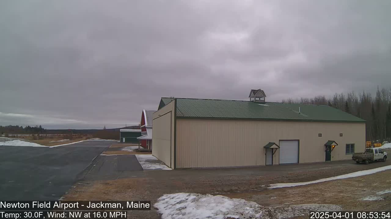 A large tan hangar with a green roof sits near a paved runway on an overcast day, with a red barn visible in the distance and some patchy snow on the ground.