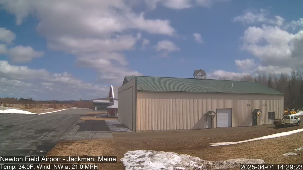 A large beige hangar with a green roof sits on an airfield under a partly cloudy sky with a northwest wind.