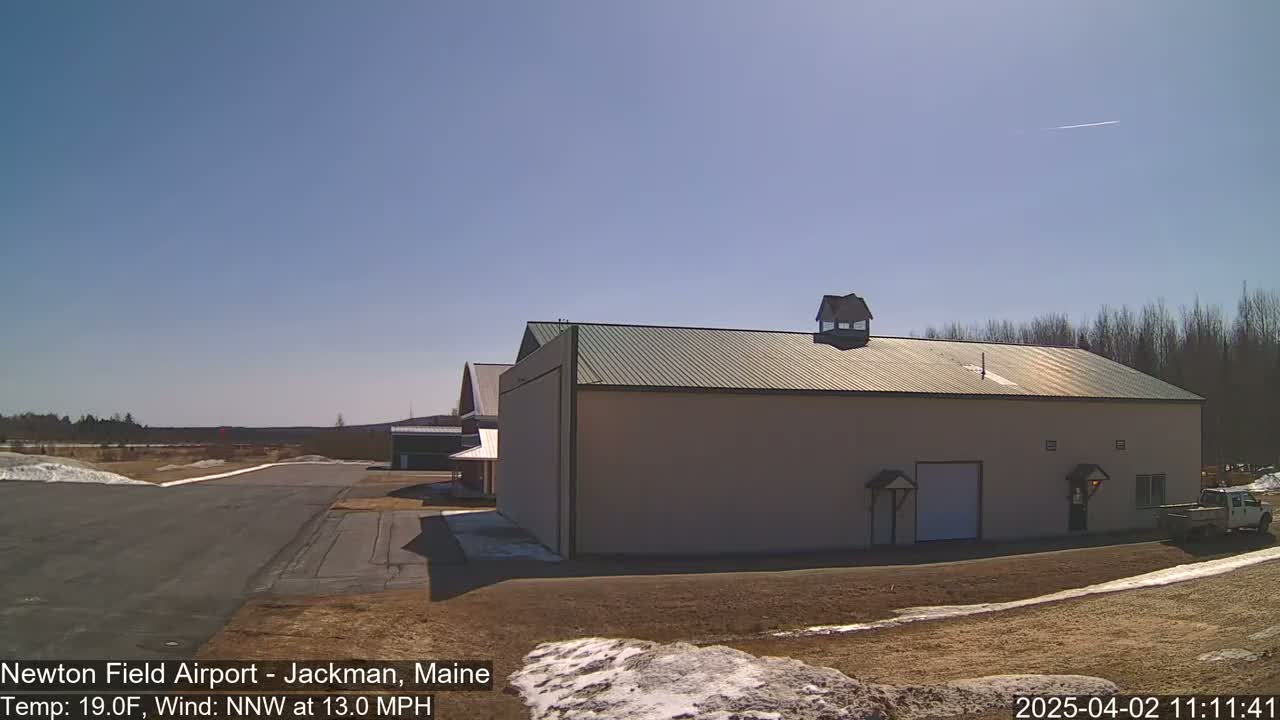 A large tan hangar with a green roof sits on an airfield under a clear blue sky, with a light dusting of snow on the ground and a few bare trees in the background.