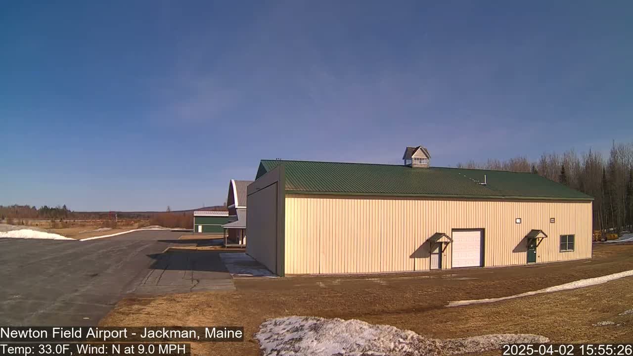 A large tan hangar with a green roof sits on a mostly clear day with a light north wind, next to a smaller red barn and a paved airport runway.