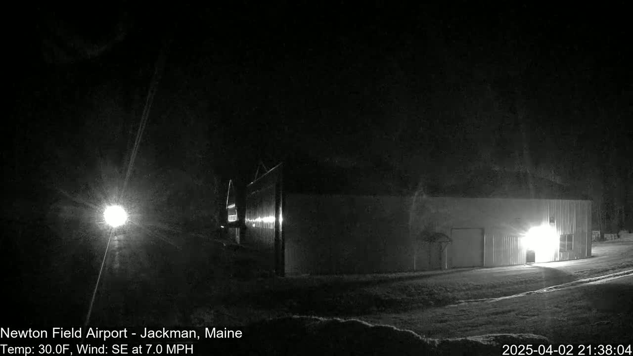 A long, low building is illuminated by a bright light at night in snowy conditions with a southeast wind.