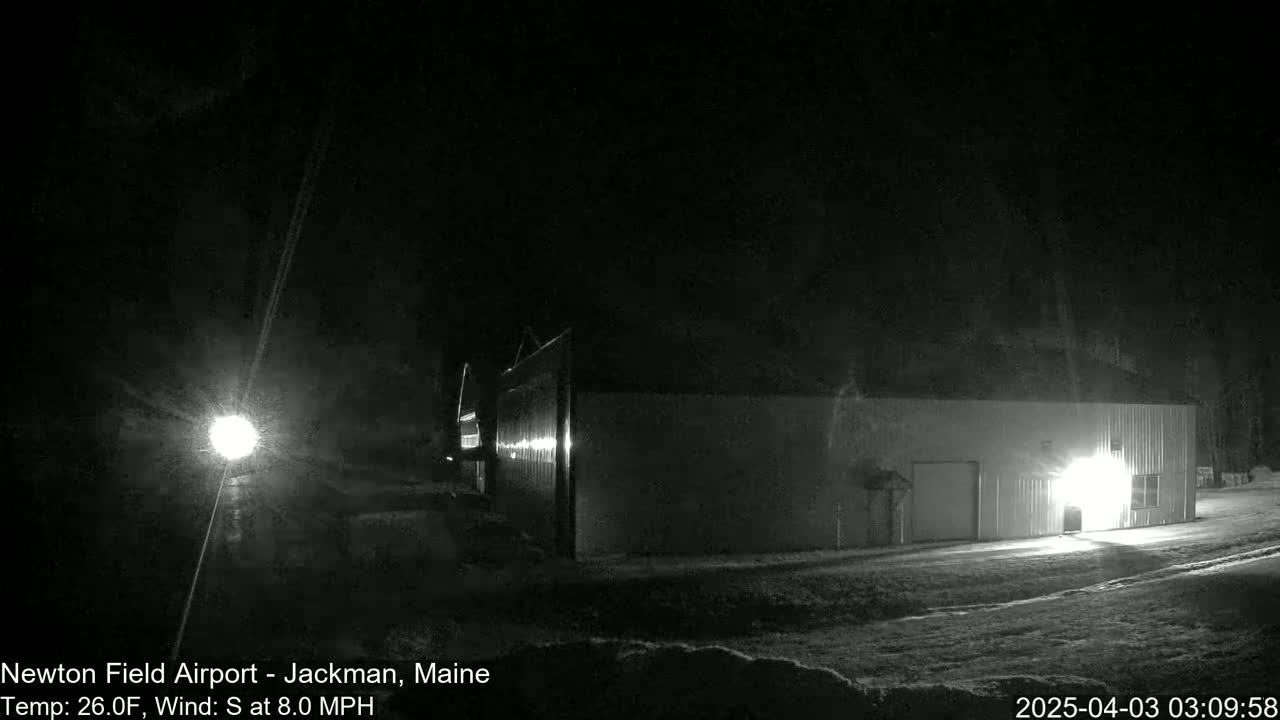 A long, low building is illuminated at night by a nearby light and its own interior lights, in snowy conditions with a south wind.