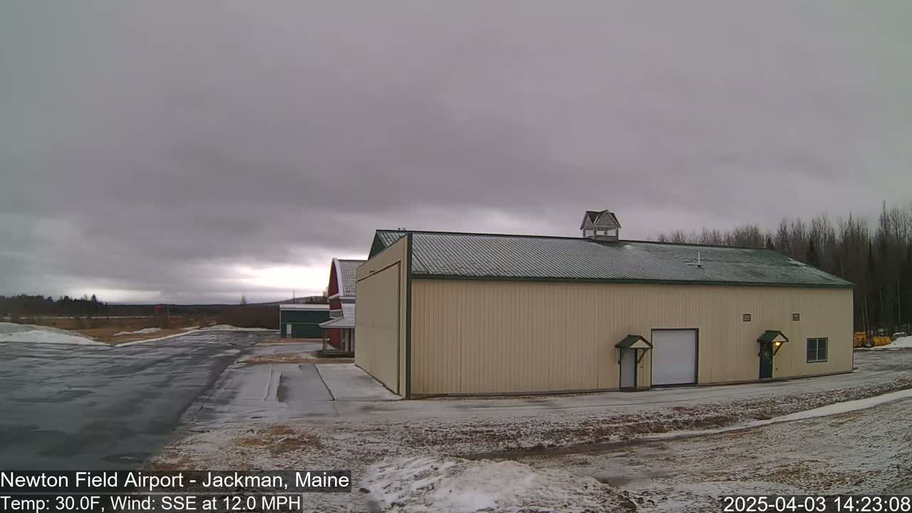 A large tan hangar with a green roof sits near a paved area and partially snow-covered ground under an overcast sky.