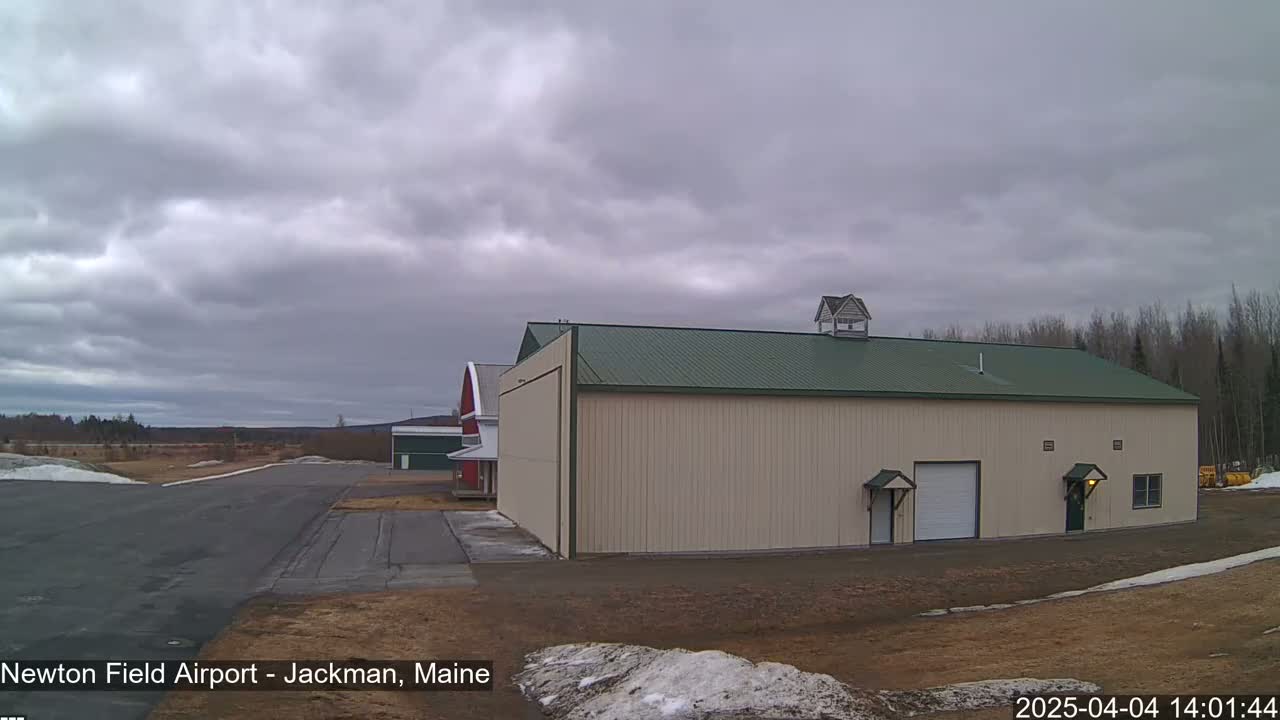 A large beige hangar with a green roof sits on an airport tarmac under an overcast sky, with a red barn visible in the background.