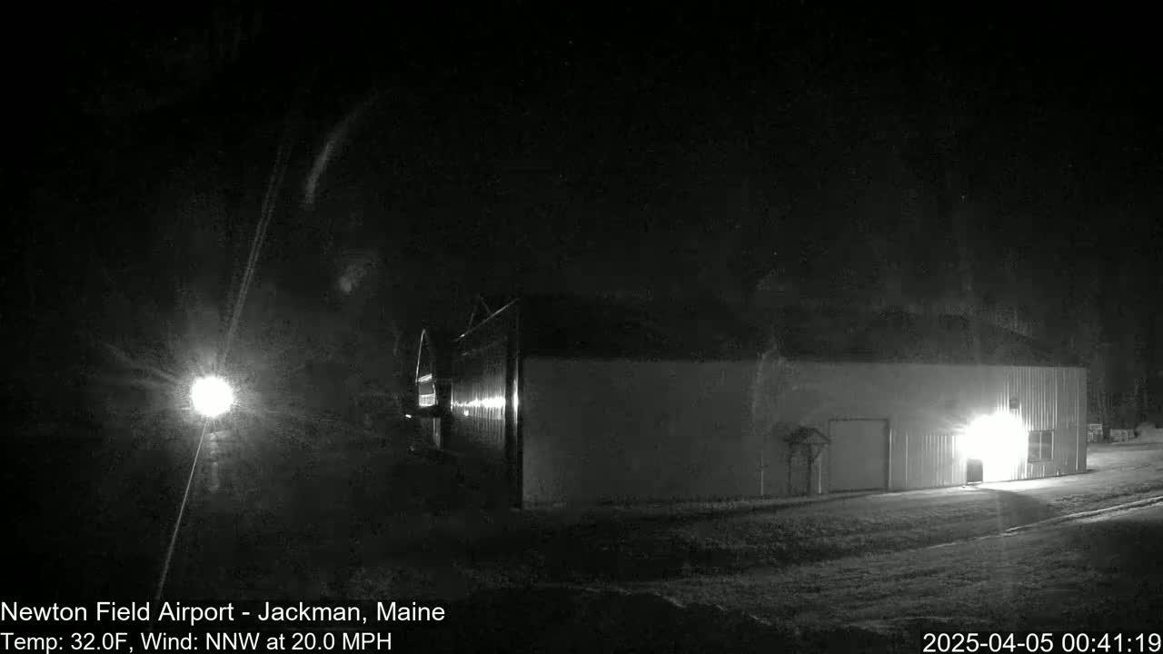 A large, light-colored building is visible at night in snowy conditions, illuminated by bright lights, with a strong north-northwest wind.