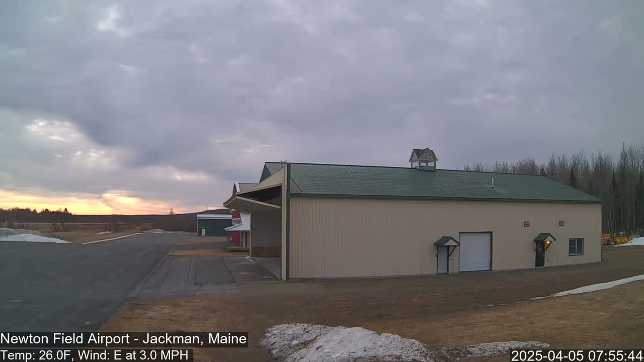 A large beige hangar with a green roof sits on an airfield under a cloudy sky with a hint of sunrise.