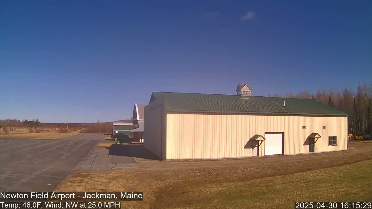 A large tan building with a green roof sits on a mostly brown airfield under a clear blue sky with a northwest wind.