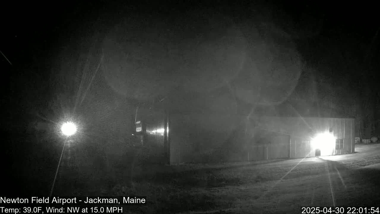 A nighttime, snowy scene shows a building illuminated by a bright light, with a strong northwest wind.