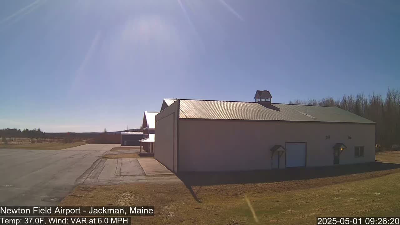 A large light-beige hangar sits on an airfield under a bright, sunny sky.