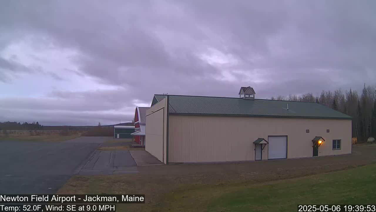 A large beige hangar with a green roof sits on an airfield under an overcast sky.