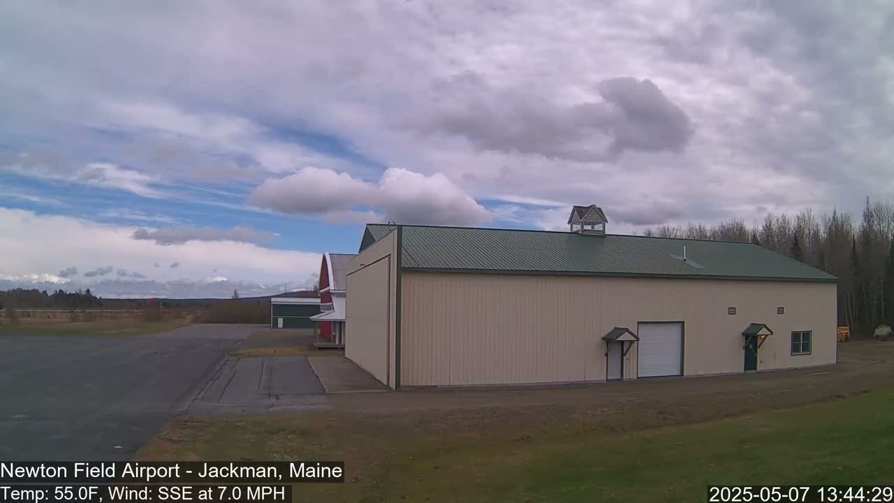 A large tan hangar with a green roof sits on an airfield under a partly cloudy sky.