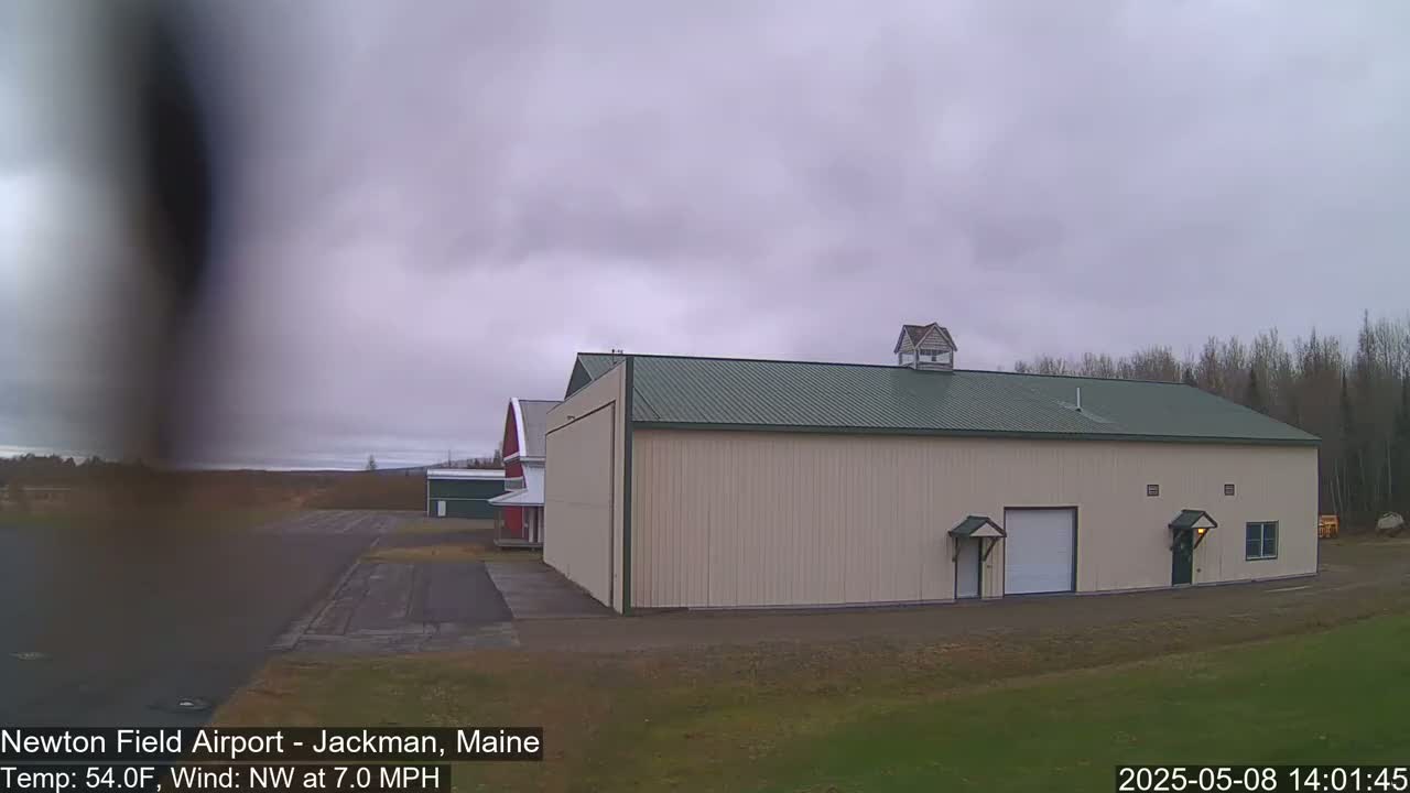 A large tan hangar with a green roof sits on an airfield under an overcast sky, with a smaller red barn visible in the background.