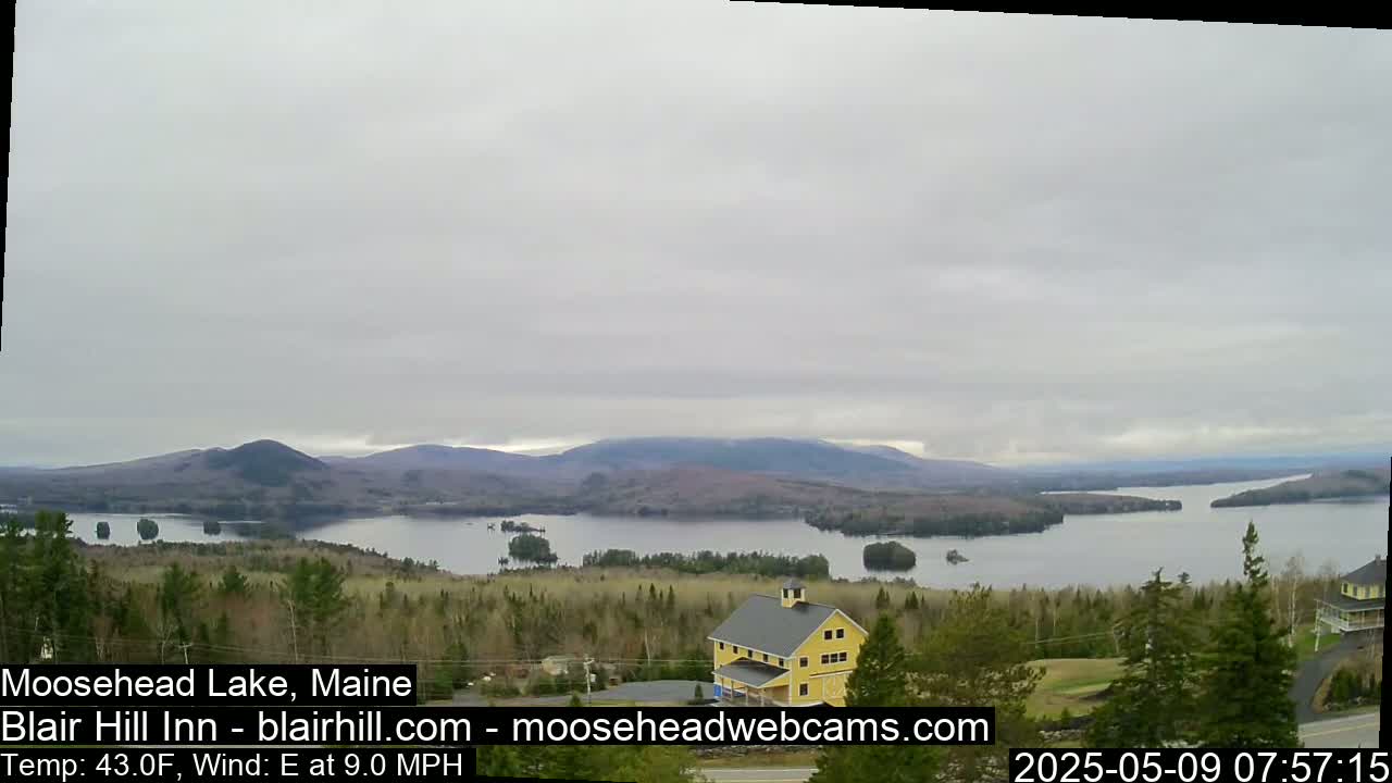 A yellow building sits on a hill overlooking a large lake and distant mountains under an overcast sky.