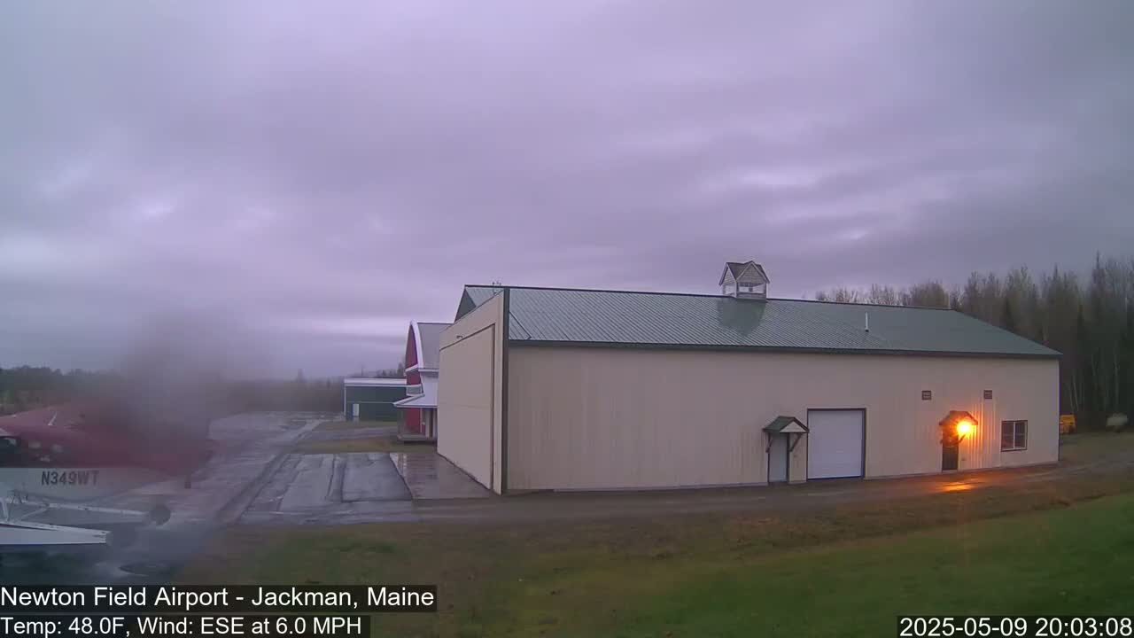 A large tan hangar sits on an airfield under an overcast sky, with a small airplane and other buildings visible in the distance.