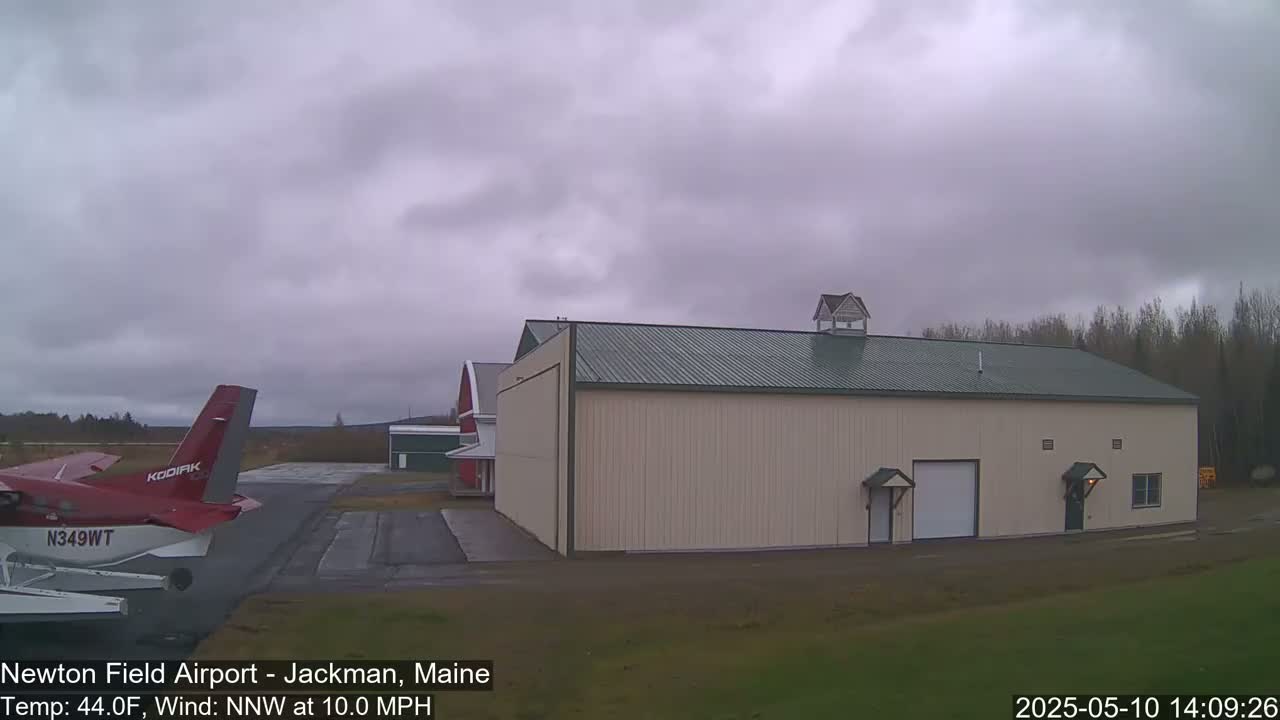 A red airplane is parked on an airfield next to a large tan building under an overcast sky.