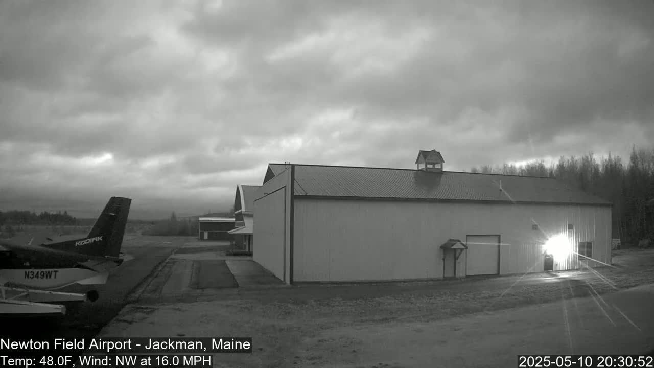 A small airplane sits on a tarmac next to a large hangar under a cloudy, overcast sky.