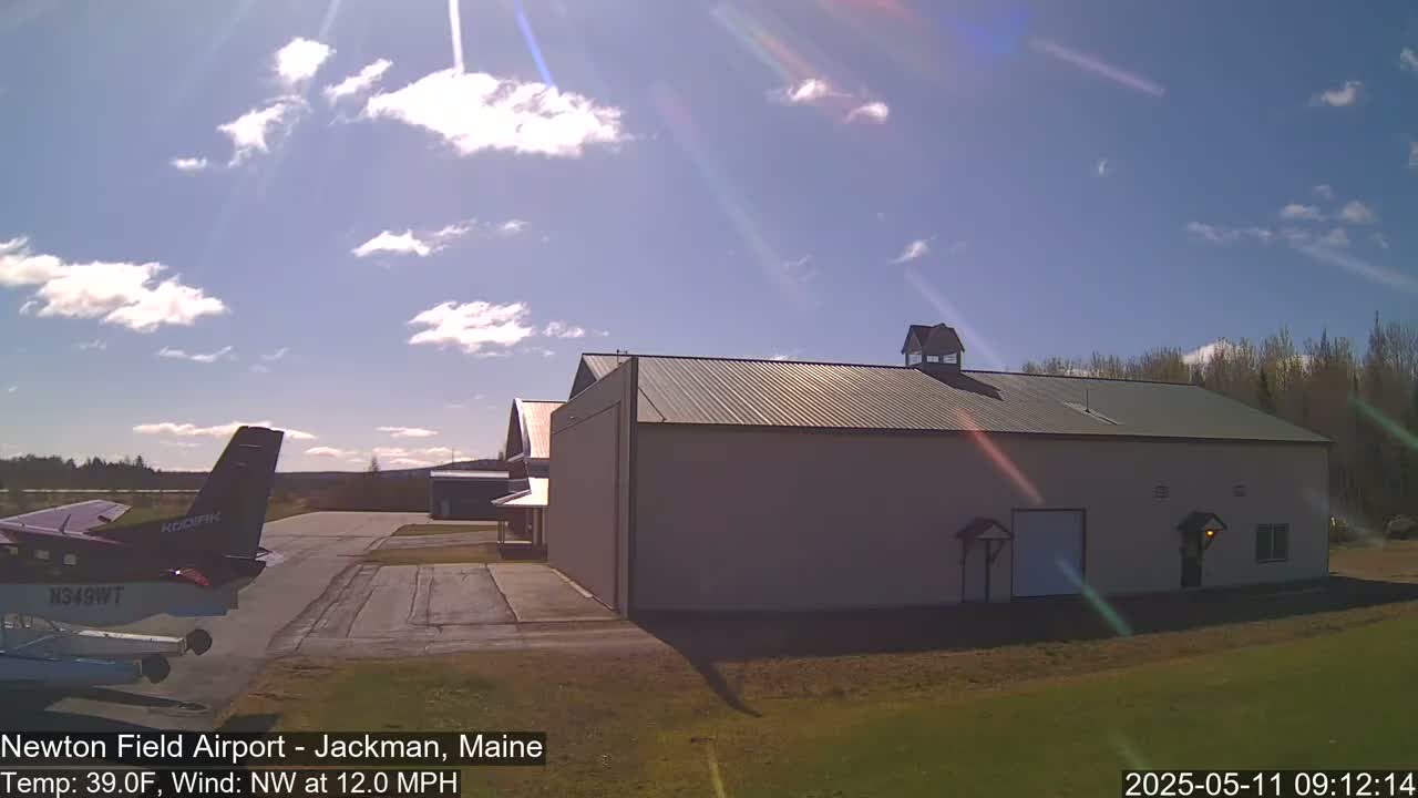 A small airplane is parked on an airfield next to a large hangar under a partly sunny sky.