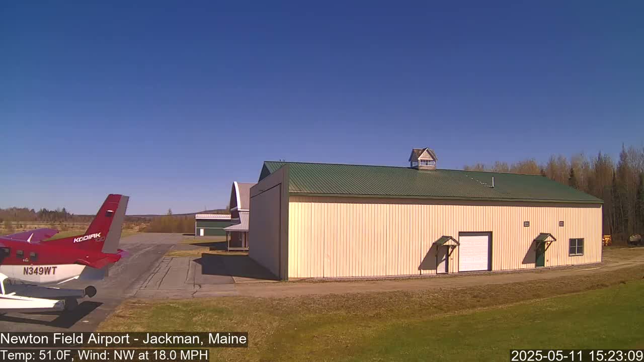 A red airplane is parked on a tarmac next to a large tan building with a green roof under a clear blue sky.