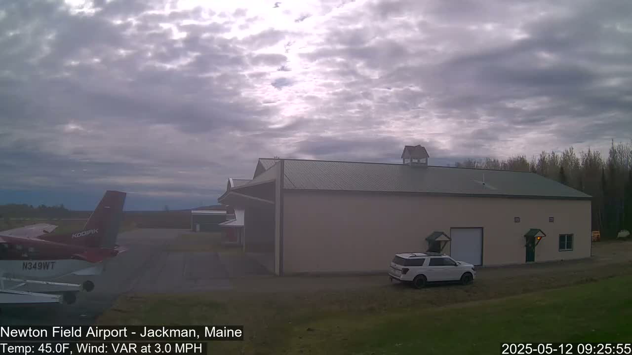 A small airplane is parked on an airfield next to a large hangar and a white SUV, under a partly cloudy sky.