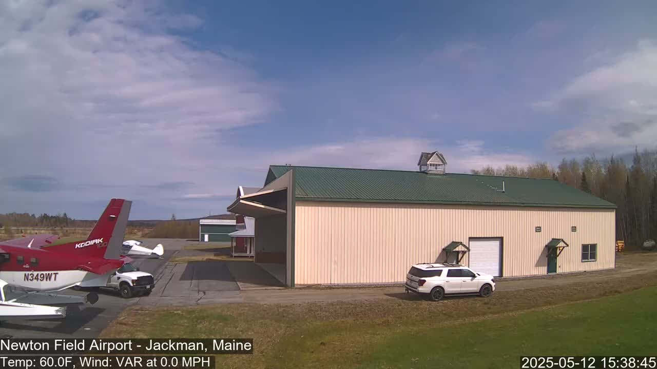 A red and white airplane is parked next to a beige hangar with a green roof at an airport under a mostly clear, bright sky.