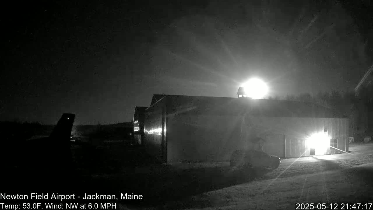 A large, dark hangar is illuminated at night by bright lights, with a vehicle parked nearby under a partly cloudy sky, and a section of an airplane is visible in the distance.