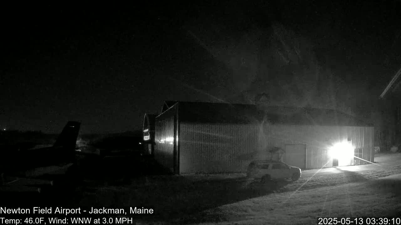 A large hangar is illuminated at night, with a vehicle parked nearby under a dark, mostly clear sky, and a small airplane visible in the distance.