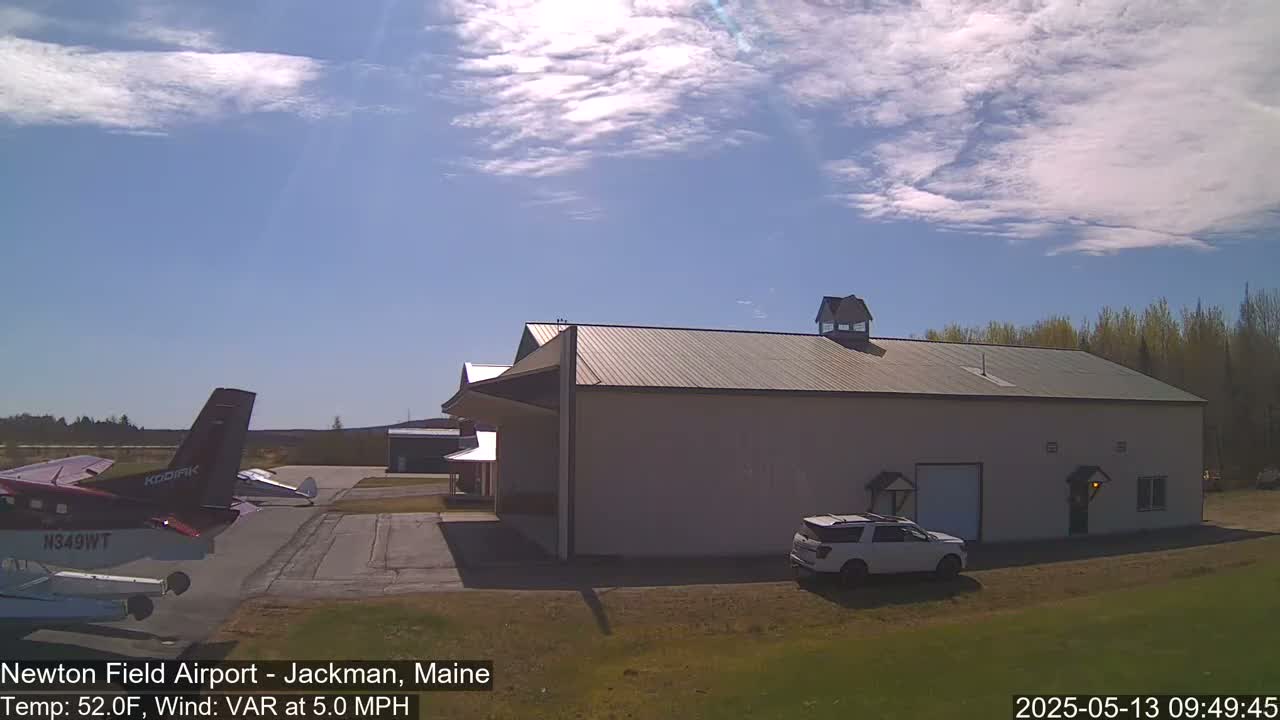 A large hangar sits on an airfield under a partly cloudy sky, with a small plane and an SUV parked nearby.
