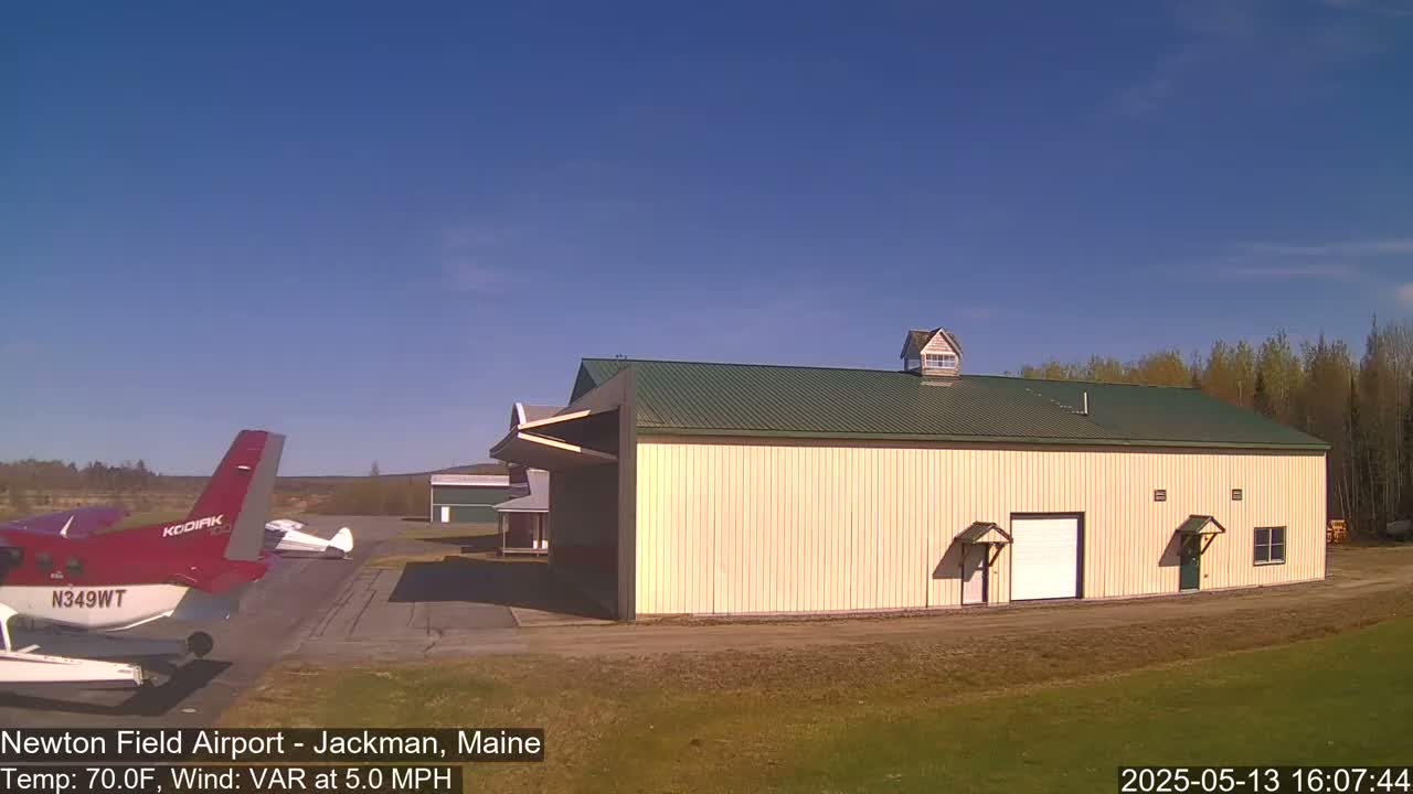 A red and white seaplane is parked on an airfield next to a large tan hangar under a clear blue sky.