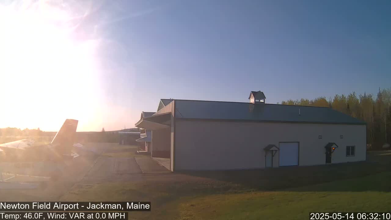 A large hangar sits on an airfield at sunrise, with a small airplane parked nearby under mostly clear skies.