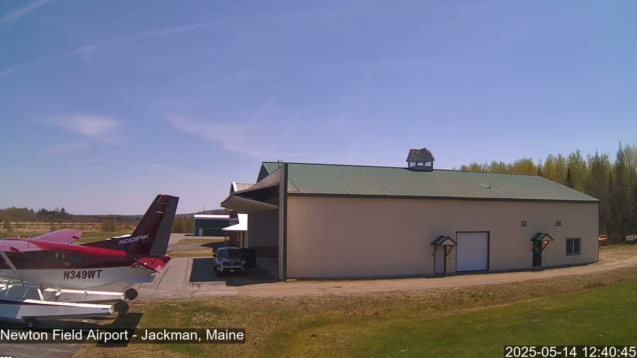 A red seaplane is parked next to a large beige hangar under a clear blue sky.