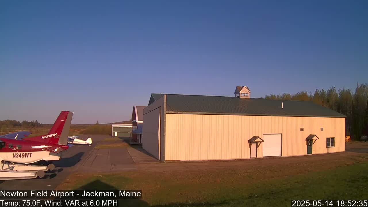 A large tan hangar with a green roof sits on an airfield next to a red airplane under a clear blue sky.