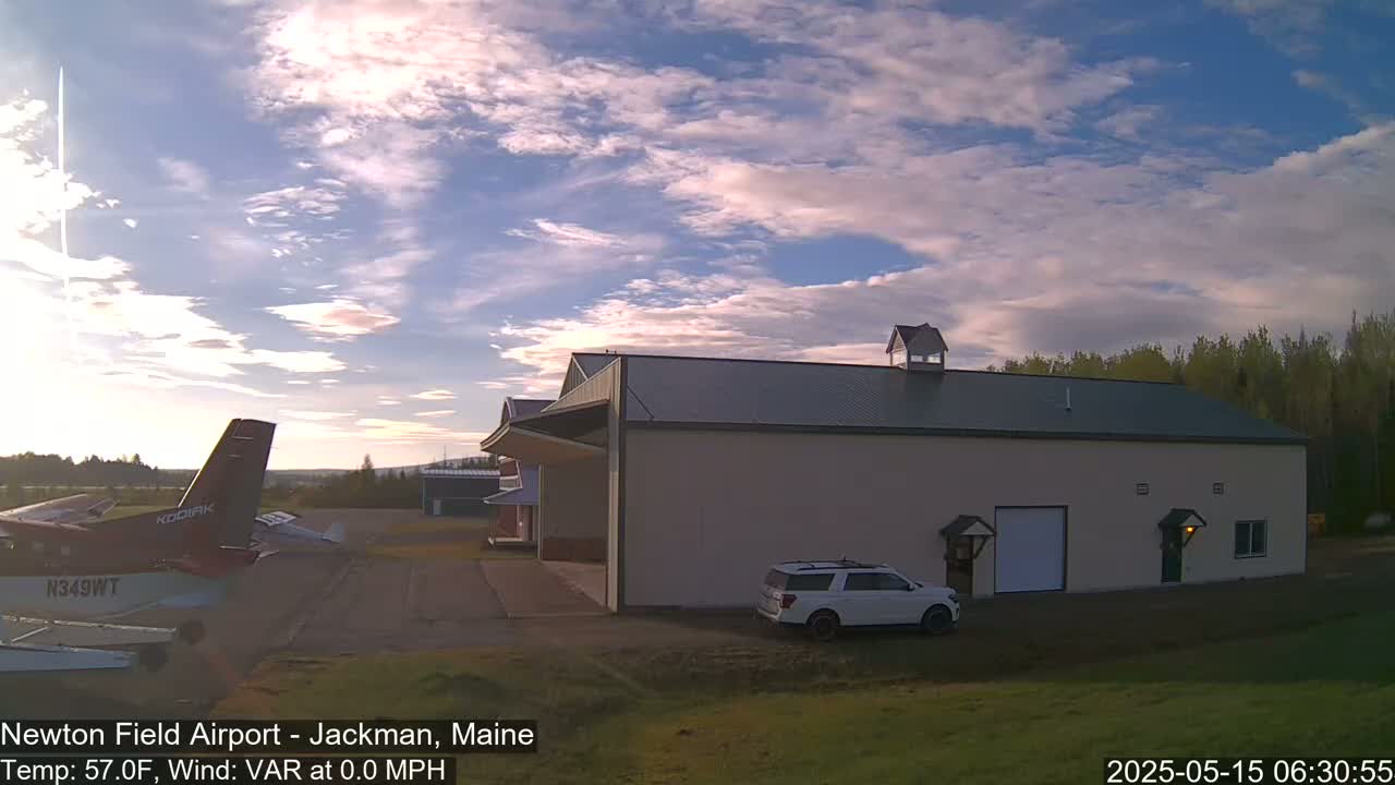 A small airplane is parked on an airfield next to a large building under a partly cloudy sky at sunrise.
