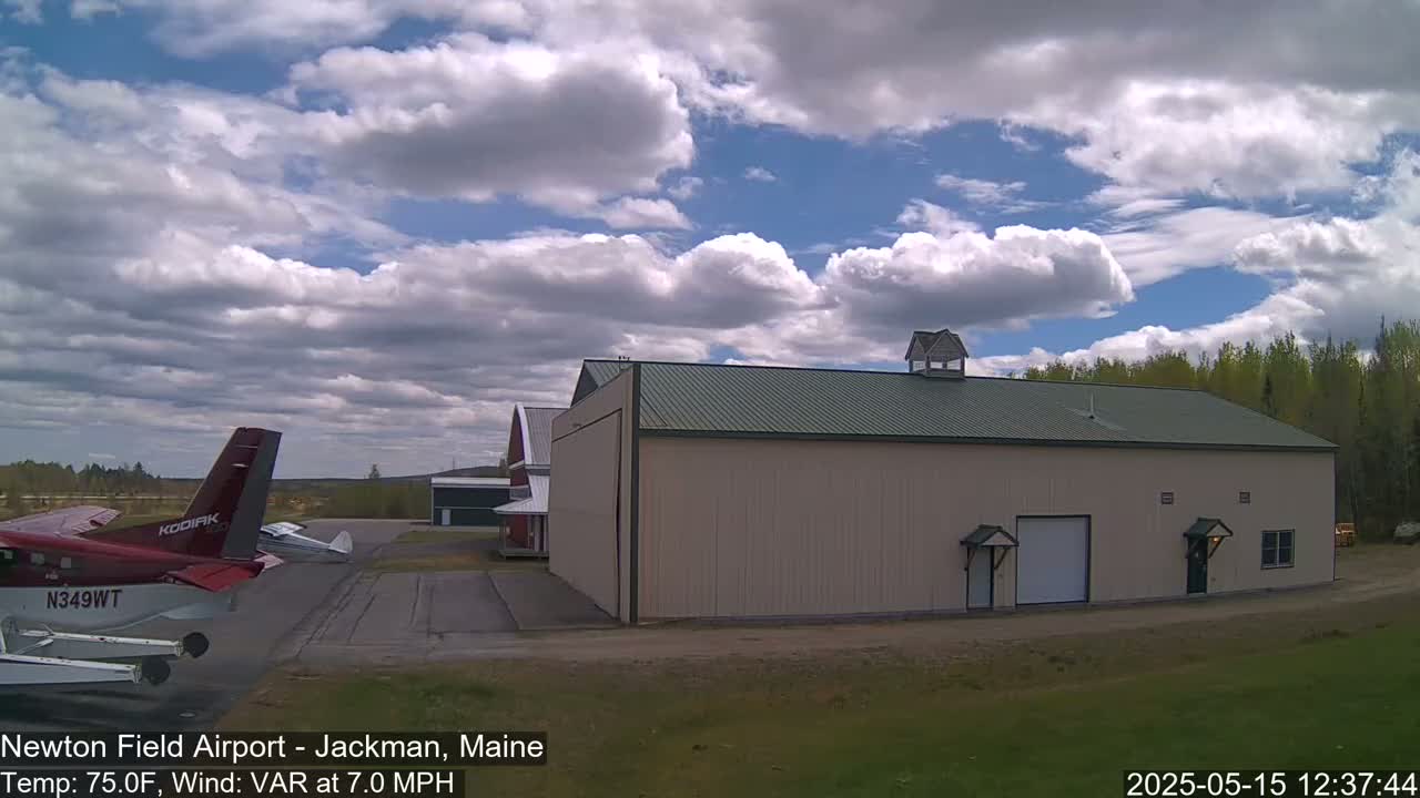 A red airplane sits on a tarmac next to a large beige hangar under a partly cloudy sky.