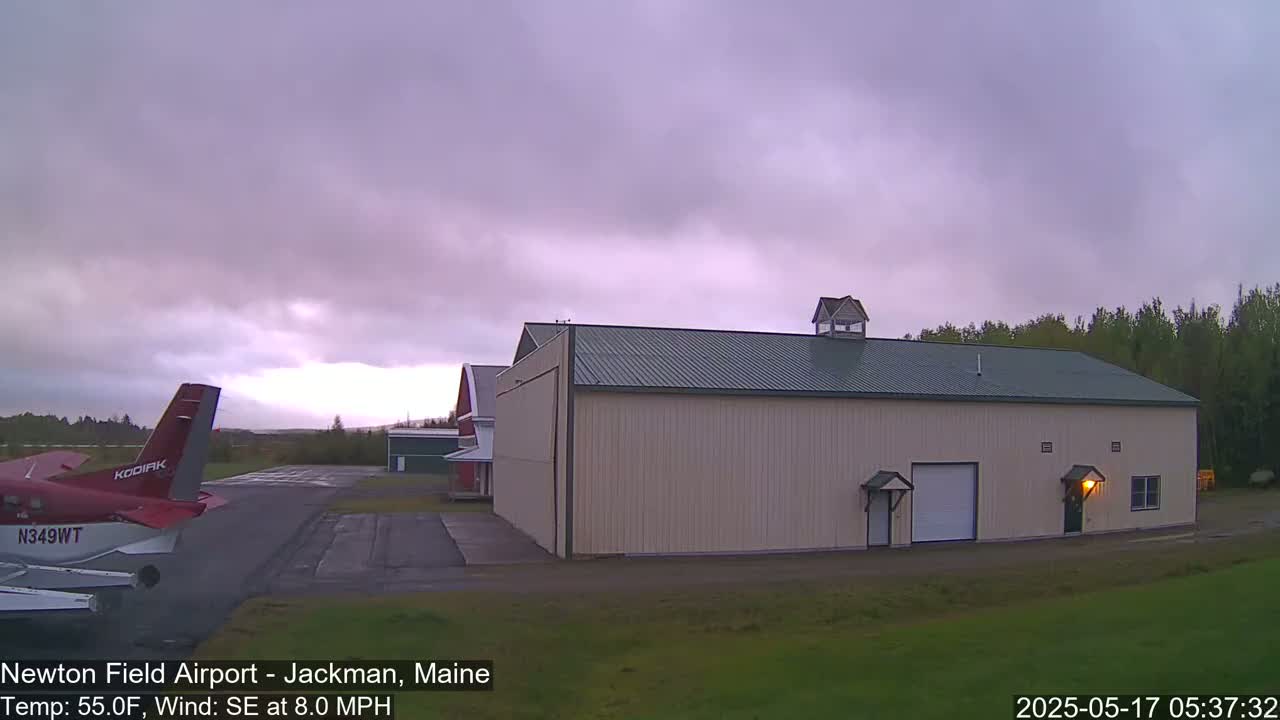 A small airplane sits on an airfield next to a large beige hangar under an overcast sky.