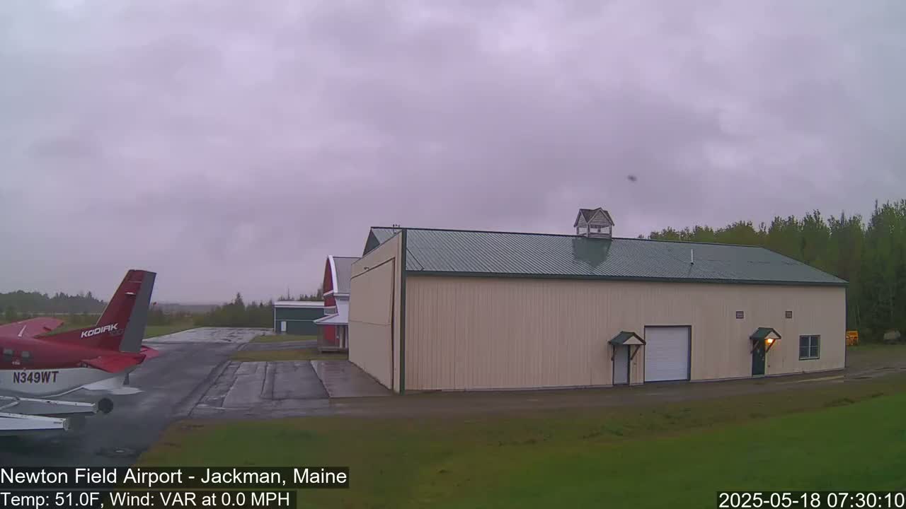 A red airplane is parked on an airport tarmac next to a large beige hangar under an overcast sky.