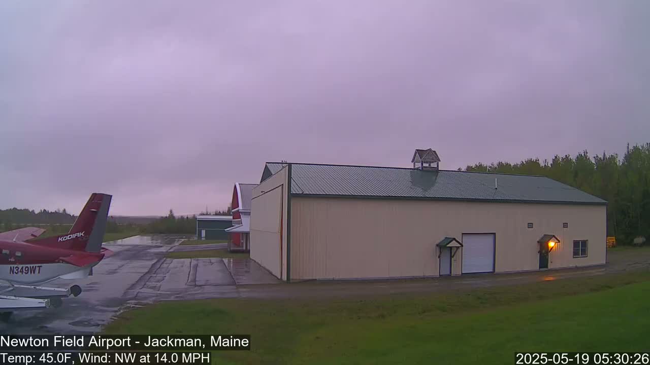 A red airplane is parked on a wet tarmac next to a large beige hangar under an overcast sky.