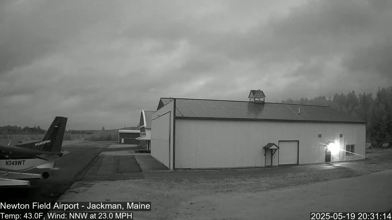 A small airplane sits on a tarmac next to a large hangar under an overcast sky, with a strong northwest wind.