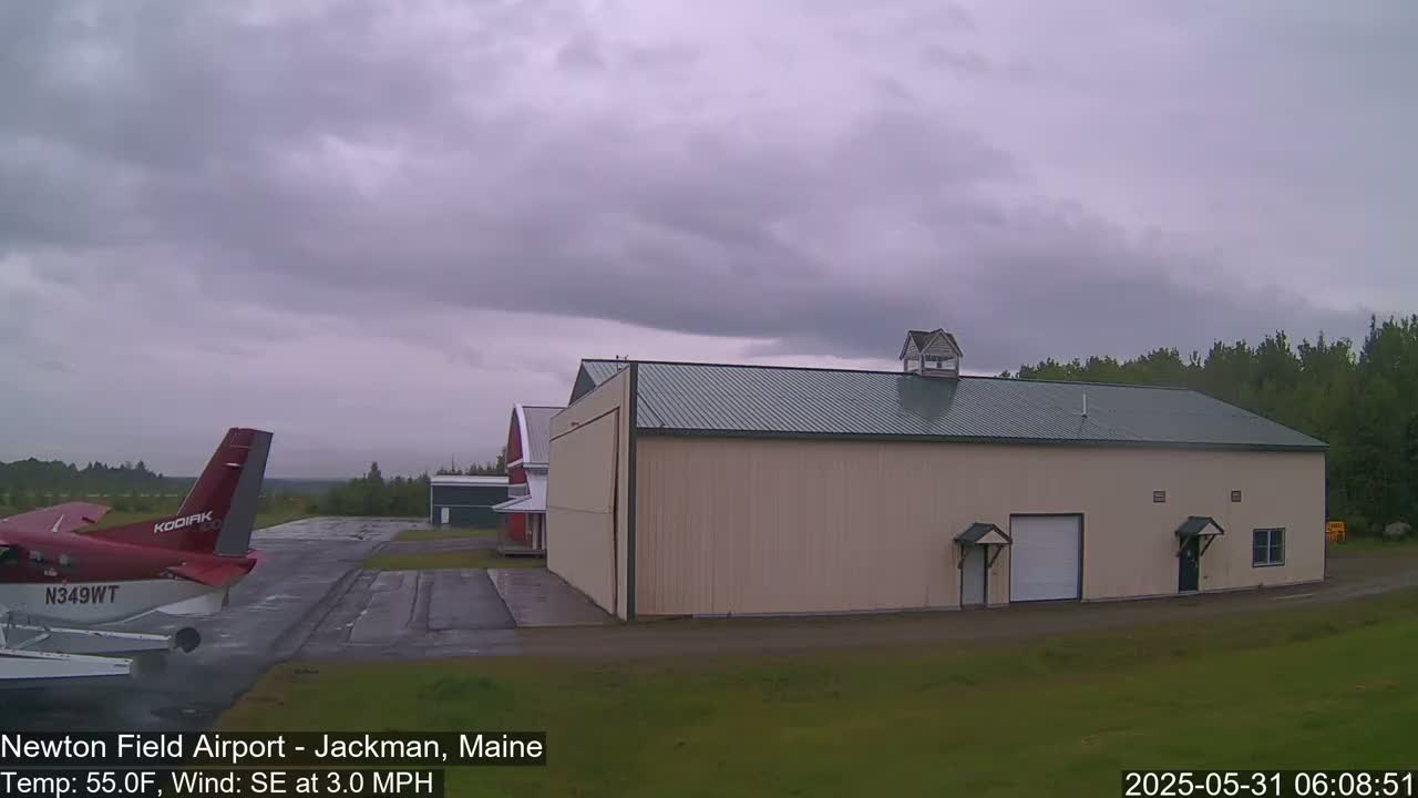 A red airplane is parked on a tarmac next to a large tan hangar under an overcast sky.
