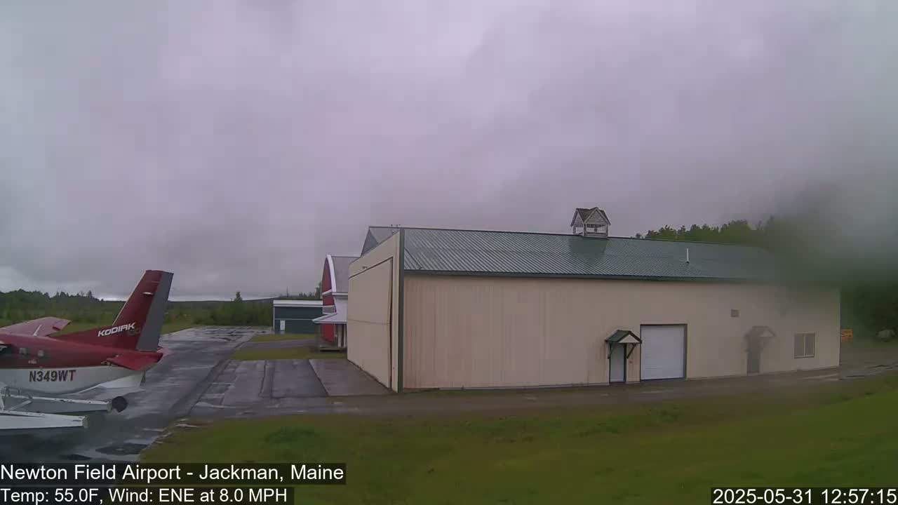 A small red airplane sits on a tarmac next to a large beige hangar under an overcast sky.