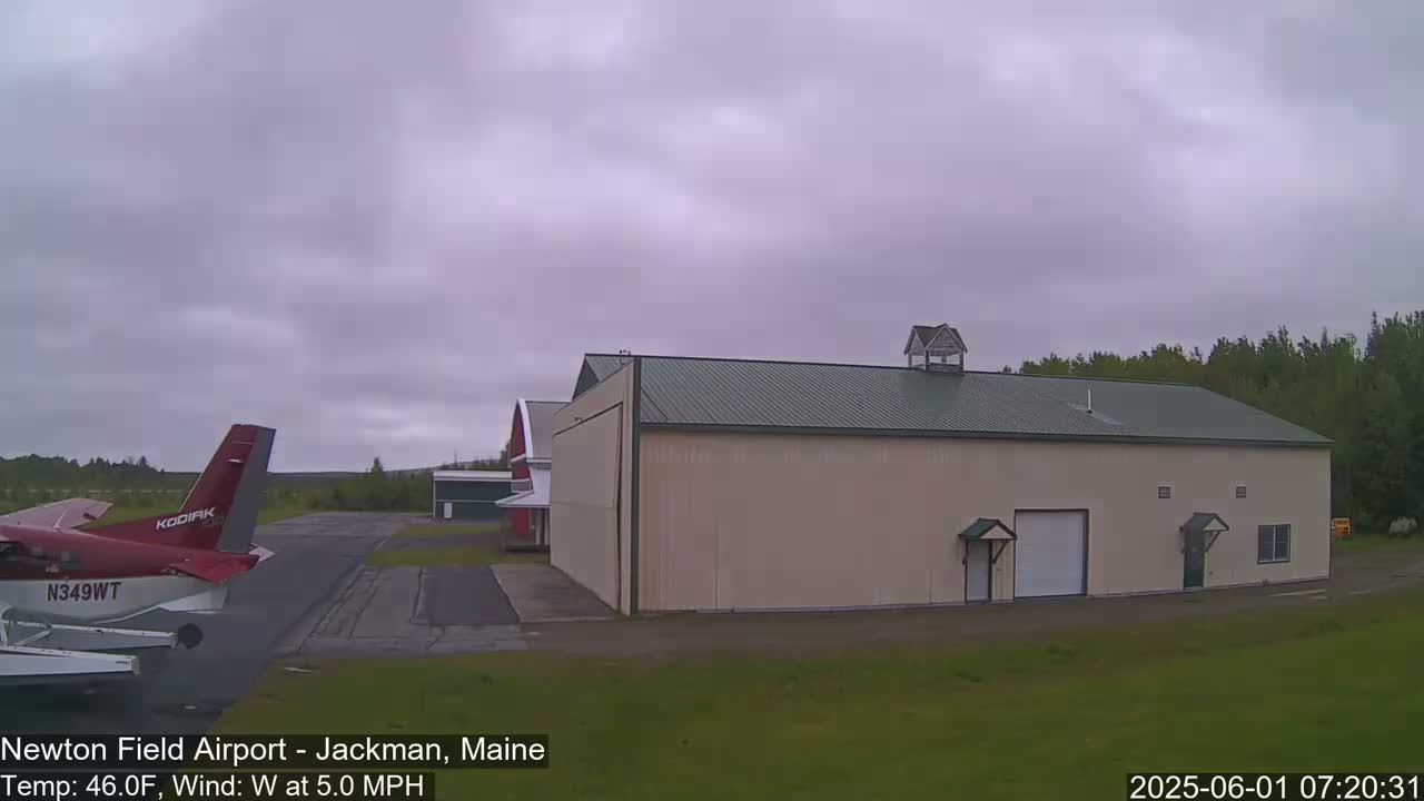 A red airplane is parked on an airfield next to a large tan hangar under an overcast sky.