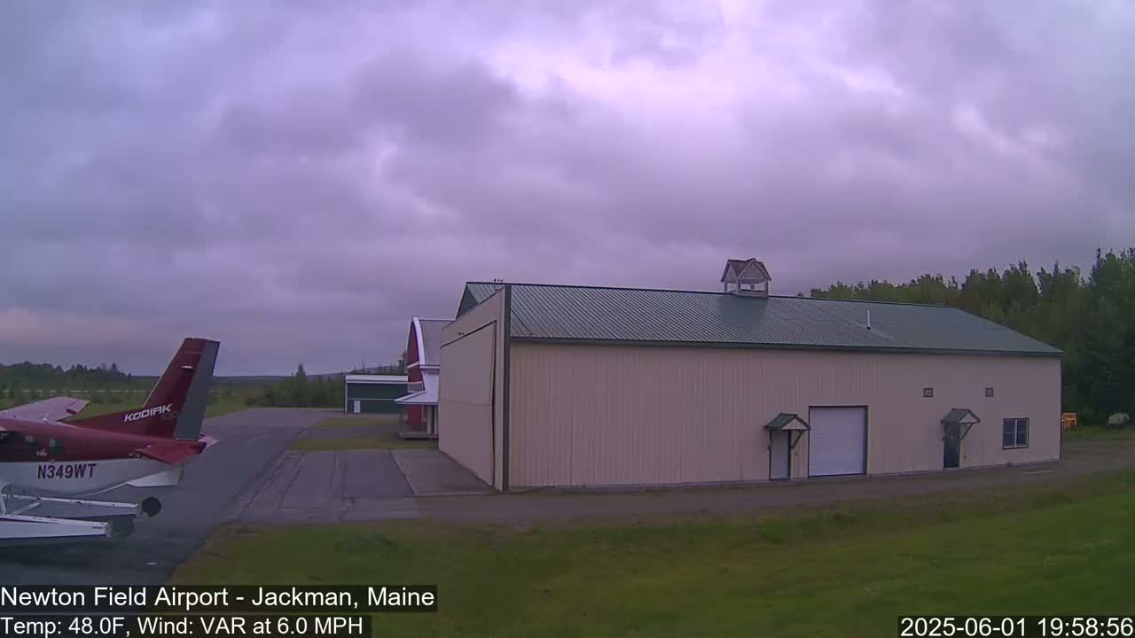 A small airplane sits on a tarmac next to a large hangar and a smaller red barn under an overcast sky.