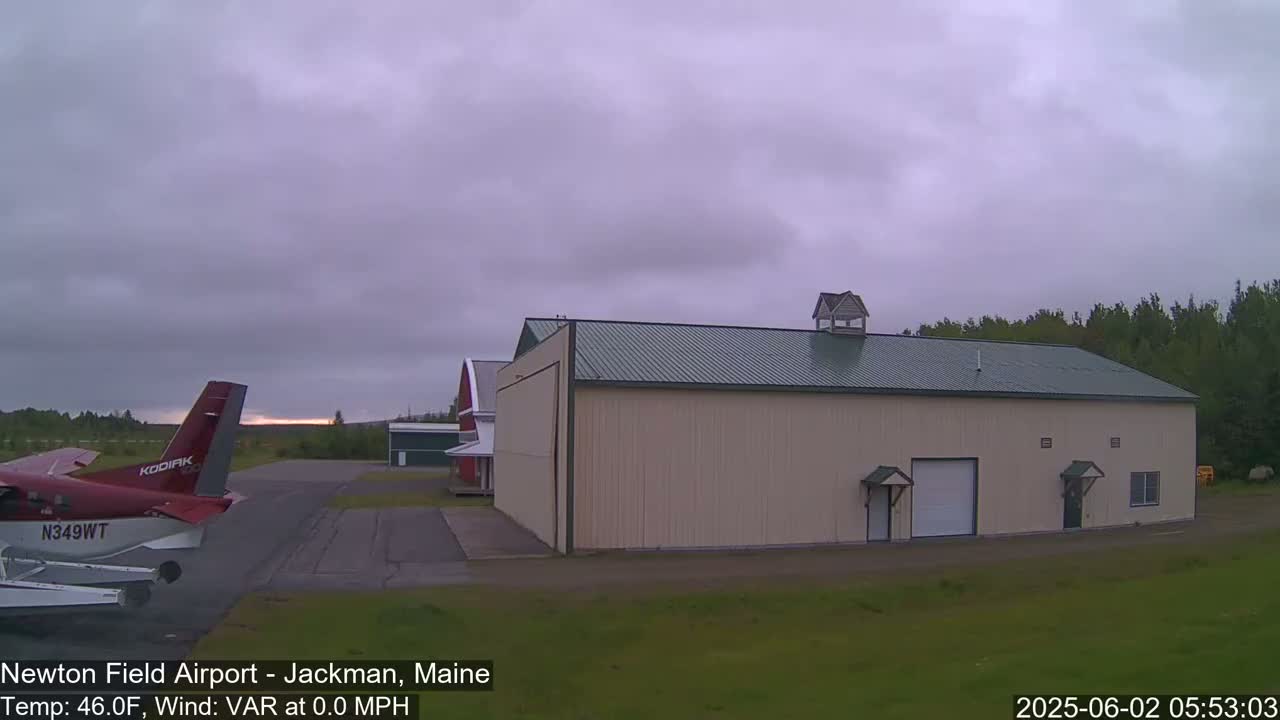 A red airplane is parked on an airfield next to a large beige hangar under an overcast sky.