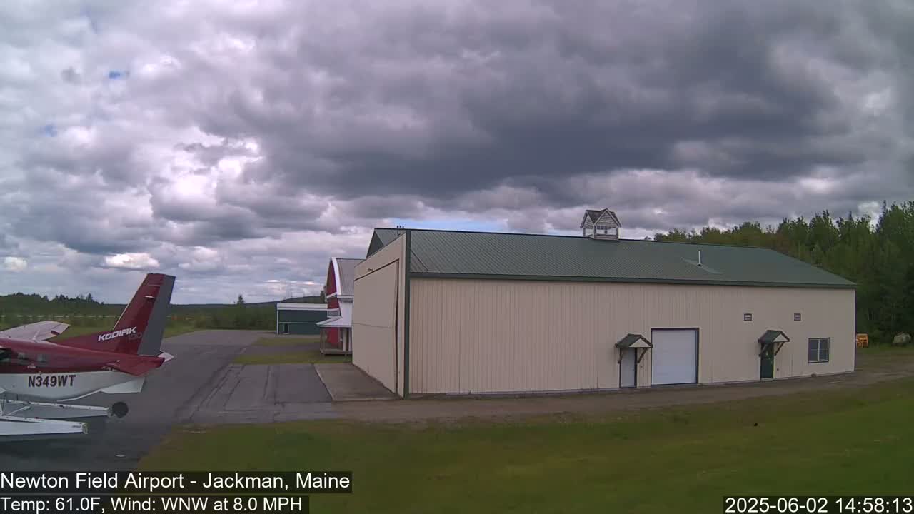 A red airplane is parked on an airport tarmac next to a large beige hangar under a mostly cloudy sky.
