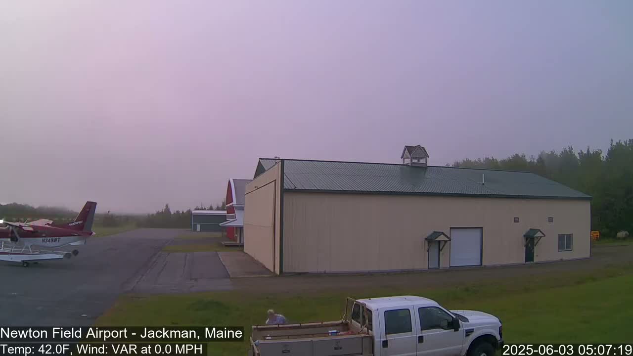 A small airplane sits on a tarmac next to a large tan hangar under a hazy, light purple sky.