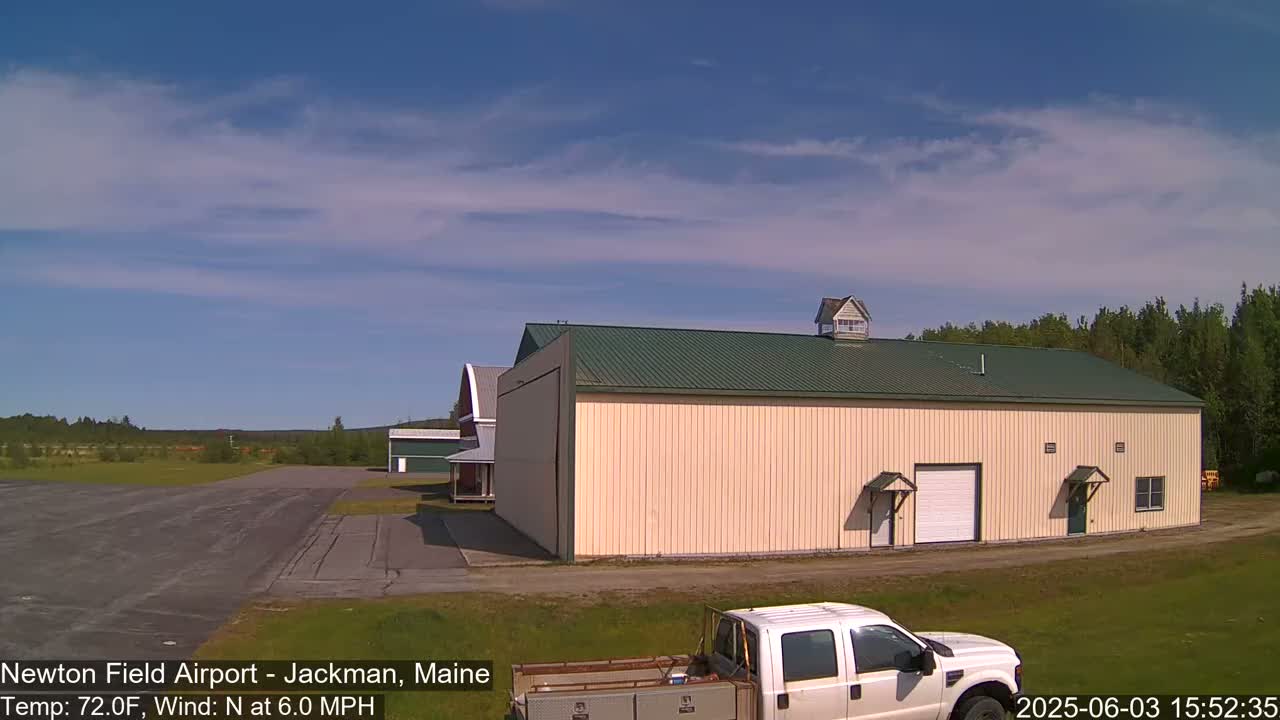 A tan hangar with a green roof sits near a runway on a sunny day with a light north wind.