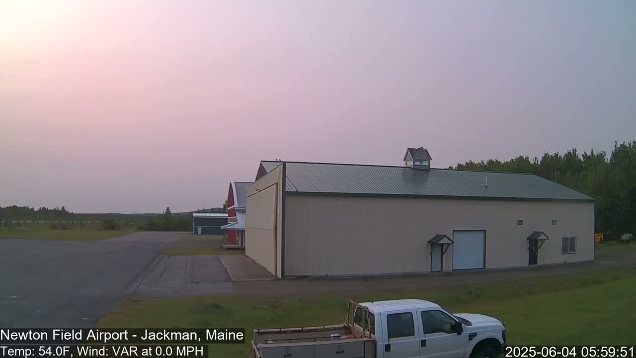 A large tan hangar sits at an airfield under a pale pink sky, with a pickup truck parked in the foreground.