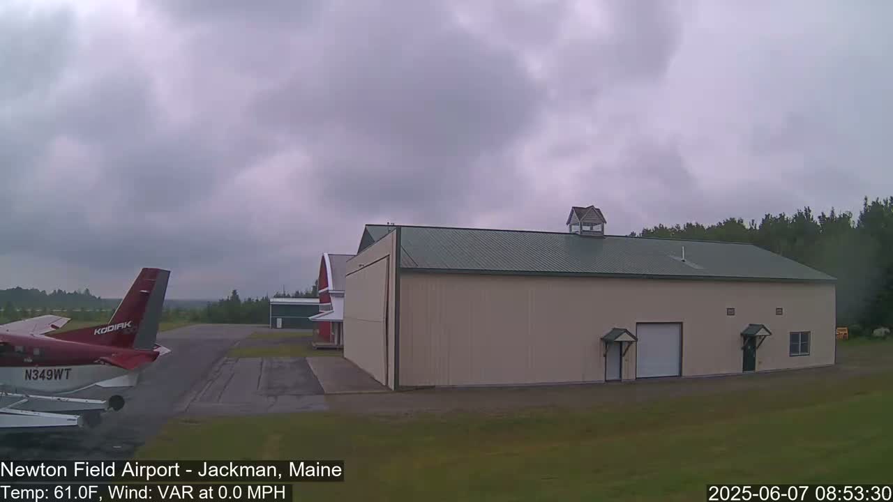 A small airplane is parked on an airfield next to a large beige hangar under an overcast sky.