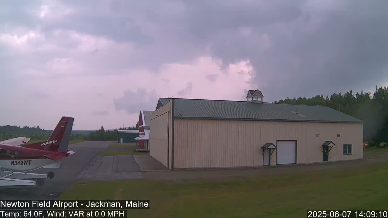 A red and white airplane is parked on an airfield next to a large tan hangar under an overcast sky.