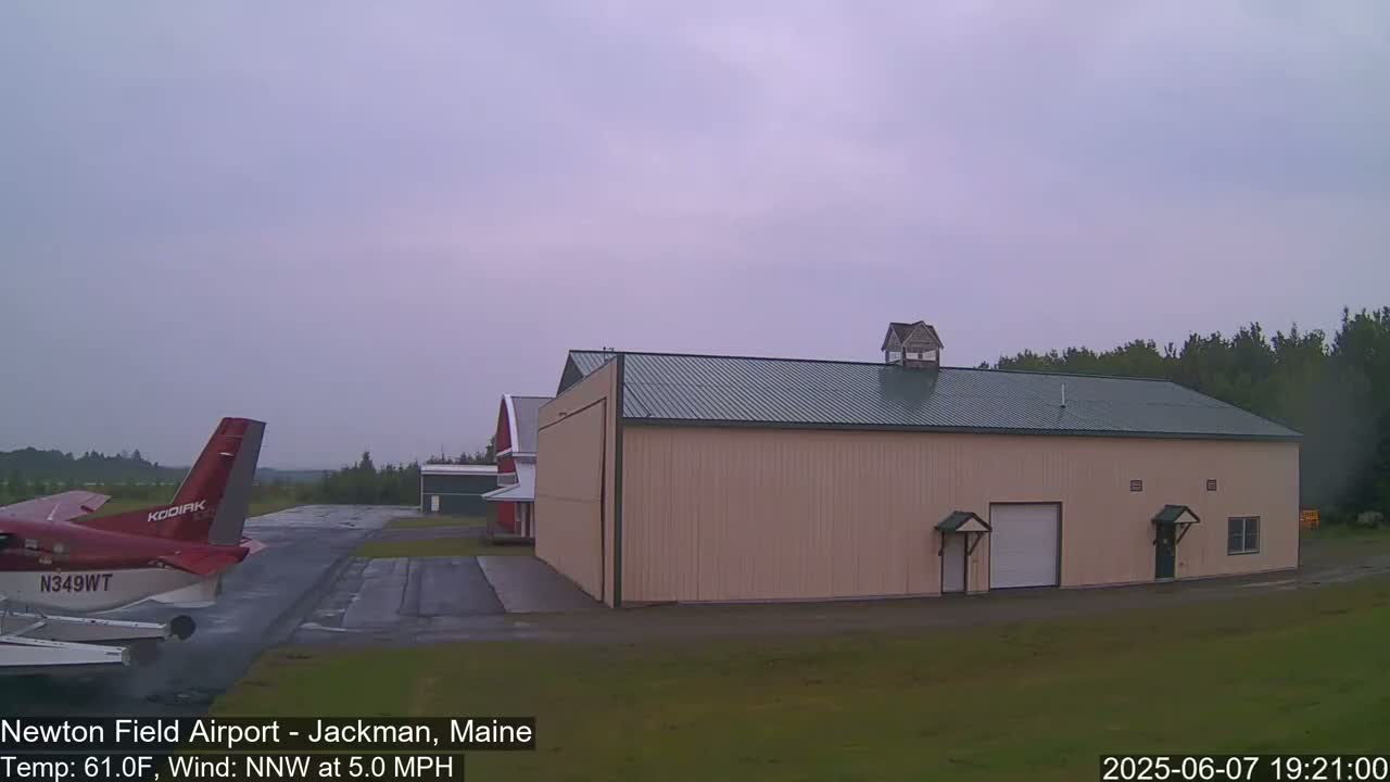 A red and white airplane sits on an airport tarmac next to a large tan hangar under an overcast sky.