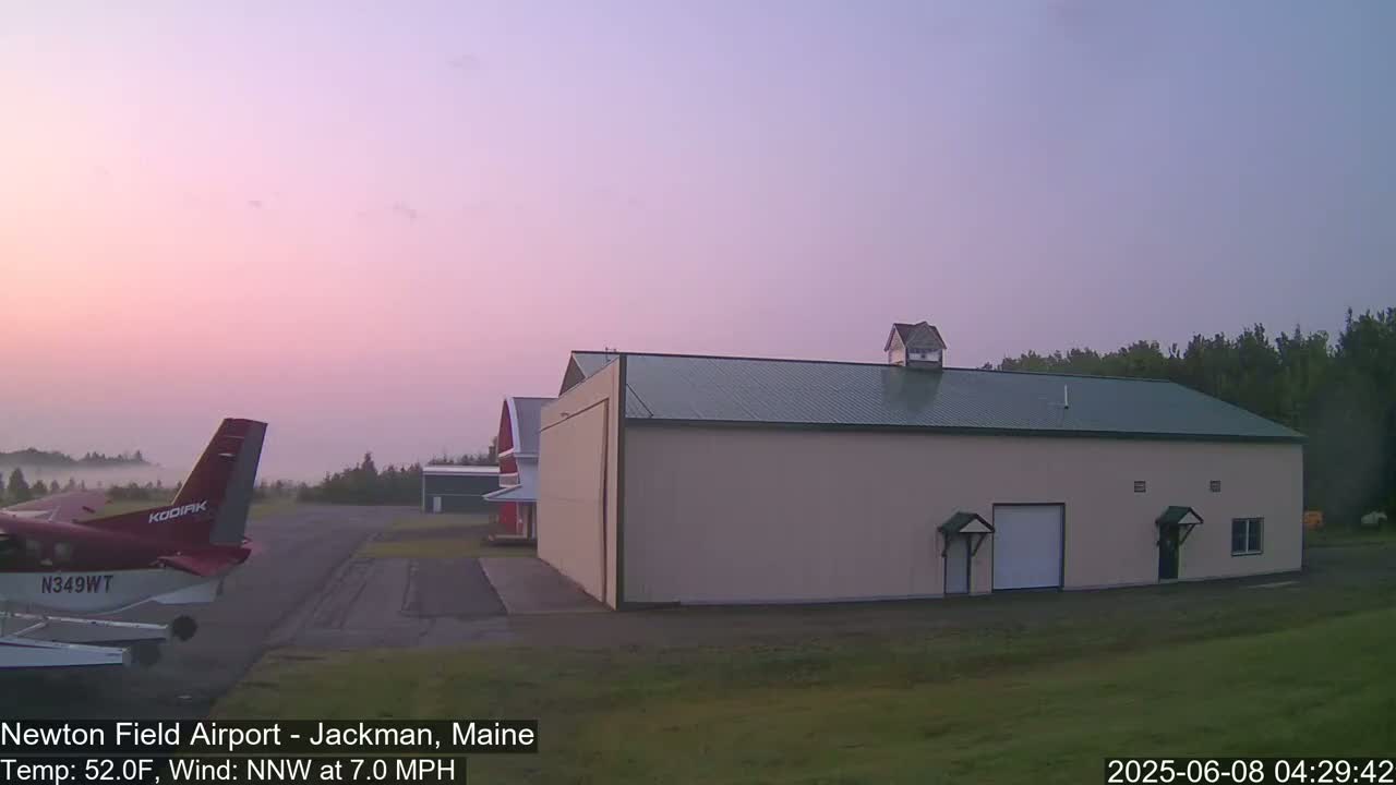 A small airplane sits on a tarmac next to a large hangar under a pale pink dawn sky with light fog in the distance.